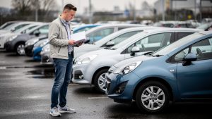 Jeune homme comparant plusieurs citadines d’occasion garées sur un parking, avec un carnet d’entretien fermé à la main.